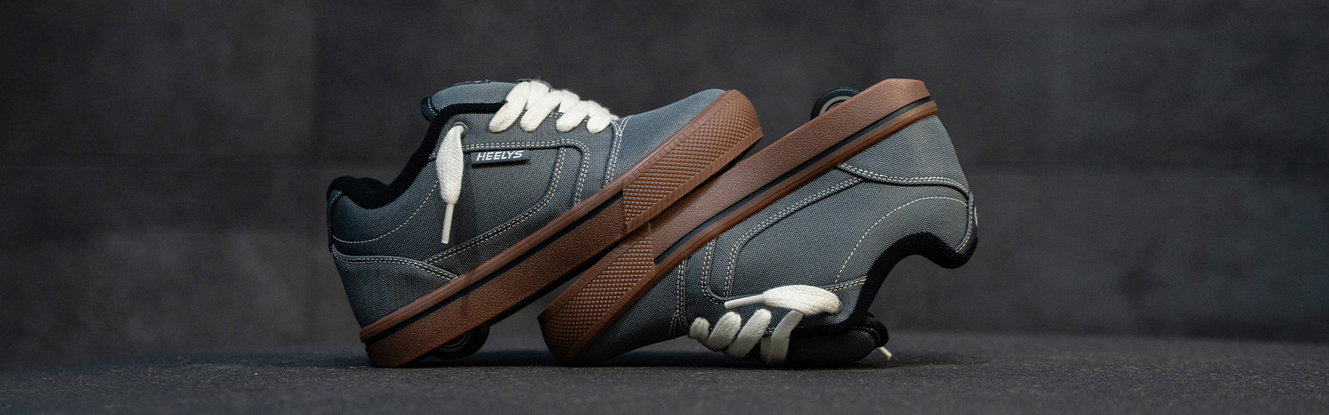 A pair of grey Pro 25 Heelys with white laces, and a thick brown sole, shown against a dark gray textured background.