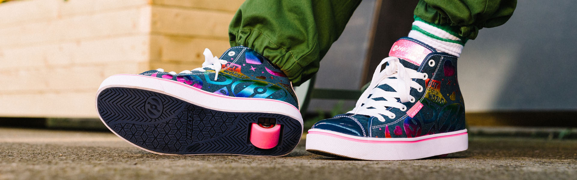 Person wearing green pants and colourful Heelys roller shoes sits on a bench next to a flower planter, with one Heelys wheel visible under the sole of the right shoe.