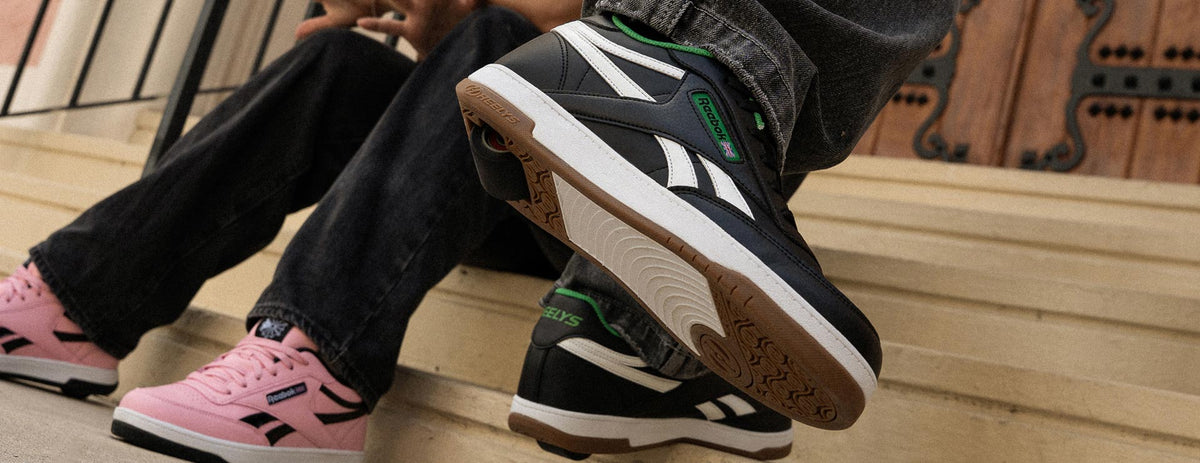 Close-up of two people seated on concrete steps, one wearing a pink Reebok Heelys and the other a black Reebok Heelys shoe with green accents.